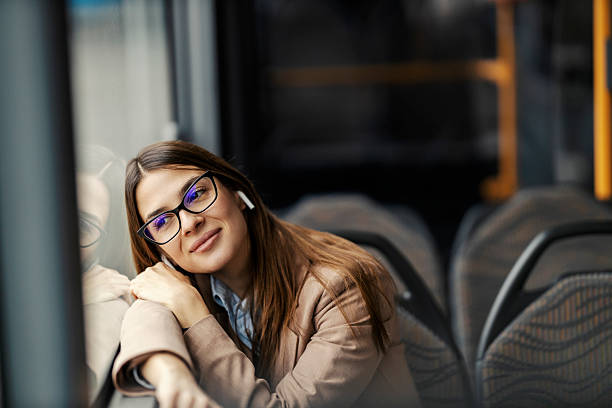 Portrait of young smiling lady leaning on bus window, listening music on headphones and looking through the window.