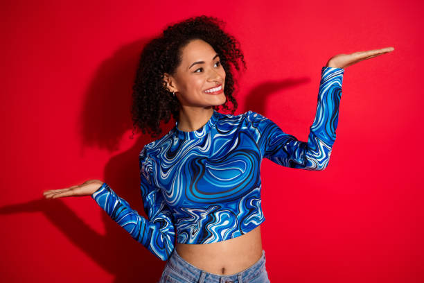 istock Smiling woman in trendy blue-patterned top presenting with raised hands against vibrant red background 2201871284