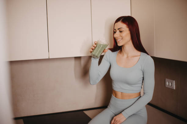 Sports woman drinking smoothie after practice, fit lady siting on the kitchen and drinking her healthy drink