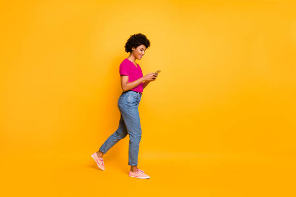 istock Side profile photo of cheerful cute attractive young influencer browsing through her telephone wearing jeans denim orange t-shirt sneakers isolated over vibrant color background 2201875279