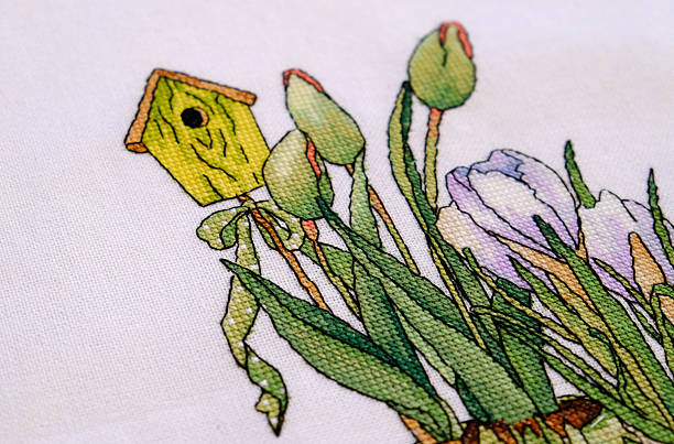 It is a handmade. The cross-stitched pattern of house is in an upright position on a grey linen background. The bobbins of color mouline are next to a tambour. These are accessories for embroidery.