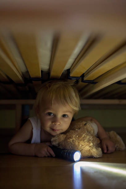 Little child, hiding under the bed, hugging teddy bear and holding flashlight, scared and sad