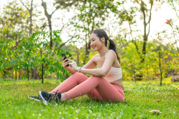 istock Young Asian Woman Sitting on Grass Using Phone After Workout Outdoors In a Park 2201877925