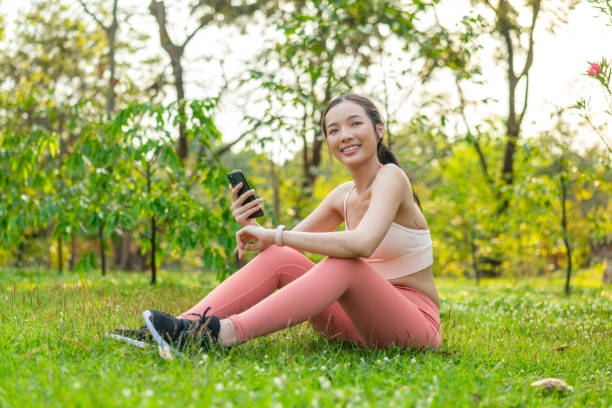 istock Young Asian Woman Sitting on Grass Using Phone After Workout Outdoors In a Park 2201877934