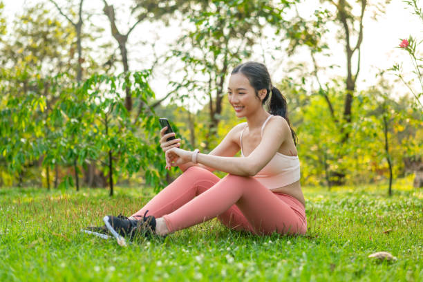istock Young Asian Woman Sitting on Grass Using Phone After Workout Outdoors In a Park 2201877966