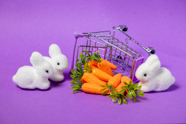 easter-themed still life with plush white bunnies, glittery artificial carrots, and a tipped-over shopping basket on a purple background