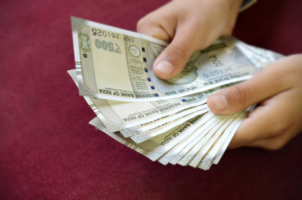 istock Two hands holding counting Indian Rupees currency paper notes cash stack of INR 500, spread or fanned out over horizontal dark maroon background 2201887616
