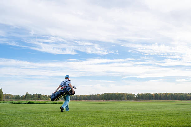 A man carrying his golf clubs and other equipment walks leisurely across a beautiful lush green golf course