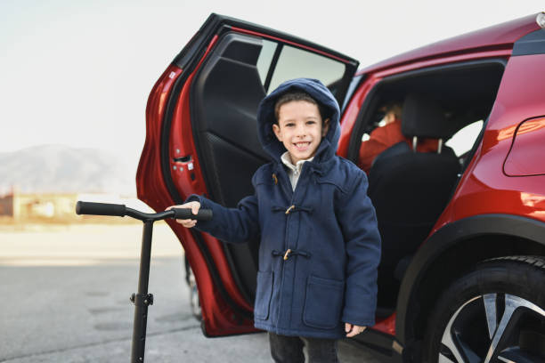 Cute Boy Posing For Camera With His Scooter While His Father is Waiting For Him in Car