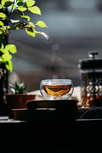 istock A transparent cup filled with tea sits on a windowsill, with visible steam rising above it. The cup is placed on books. Sunlight filters through green leaves, adding a natural, vibrant touch. 2201892915