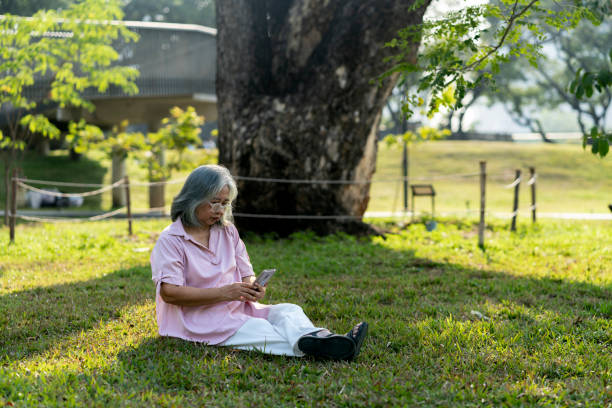 istock Senior woman using mobile phone during sitting under large tree in park 2201899363