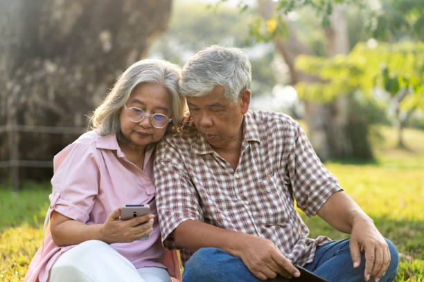 Asian elderly couple uses mobile phone while sitting on the grass under giant tree in public park on sunny light. A wheelchair parked nearby, They looking on smartphone and smile with happy together