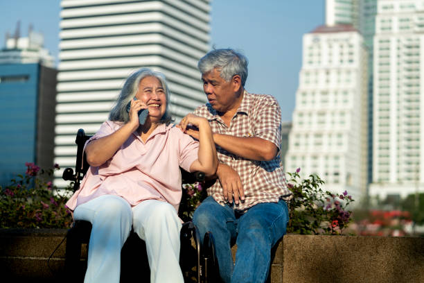 Happy senior couple sits by the waterfront at city park, Wife smiling during talking on smartphone, husband placed his arms on her shoulder, They enjoying a sunny day