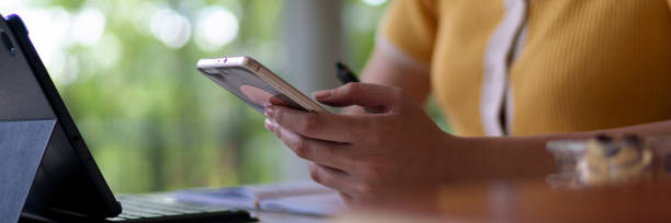 istock Young Asian woman using a smartphone while studying with a tablet in a university cafe, with books and a drink on the table 2201911144