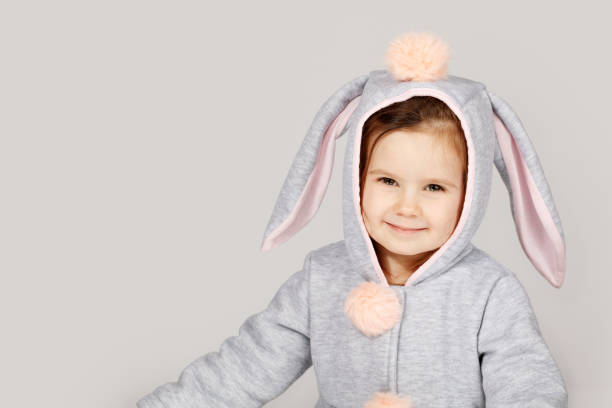 a cute girl dressed in a soft pastel bunny costume with floppy ears and a fluffy pom-pom on the hood sits and smiles against a clean background. The child expresses joy and playfulness, making the image perfect for Easter, children's themes and festive concepts