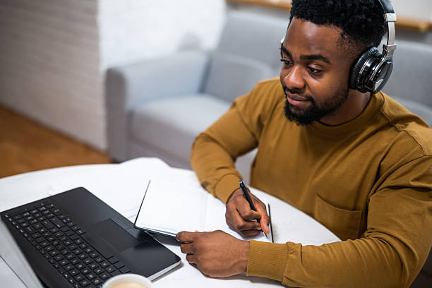 istock Young black male with headphones using laptop and taking notes at home 2201917430