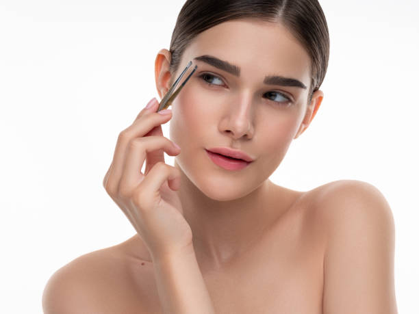 A woman carefully tending her eyebrows with tweezers, showcasing attention to self-grooming. Her focused expression emphasizes delicate care and the beauty enhancements associated with personal grooming routines.