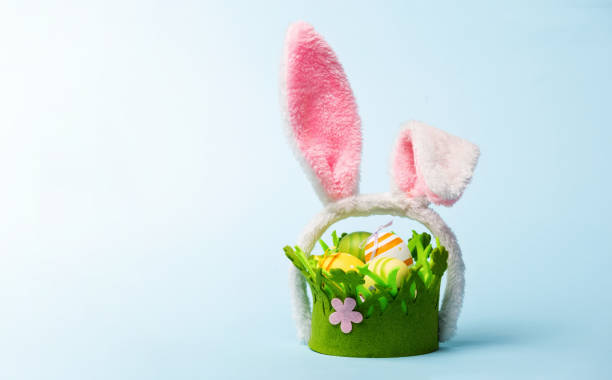 istock Funny Easter basket with decorated colored eggs. Green basket with painted eggs and pink bunny ears on a pastel blue background. 2201929280