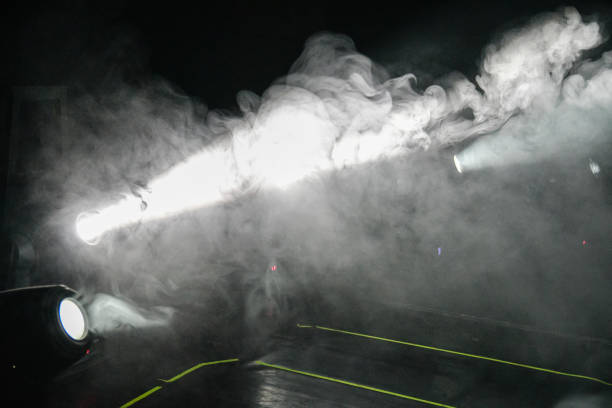 Bright spotlights cutting through swirling smoke on a concert stage, creating a moody and atmospheric lighting effect. The combination of haze and beams of light enhances the dynamic and immersive feel of a live music performance.