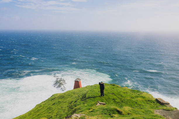 Front view of female explorer in distant staying at the edge of the cliff overlooking majestic ocean during sunny day on Faroe Islands