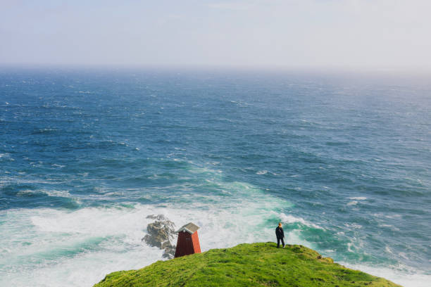 Rear view of female explorer in distant staying at the edge of the cliff overlooking majestic ocean during sunny day on Faroe Islands