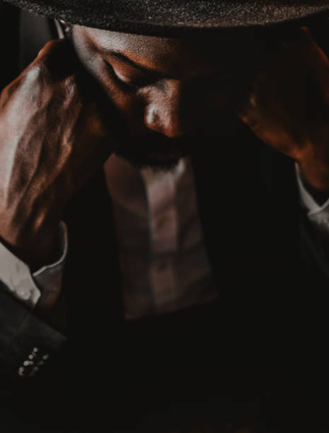 Close-up of the face of a black man with a hat. He is supporting his head with his hands. He is in a room with some daylight and is shrouded in darkness.