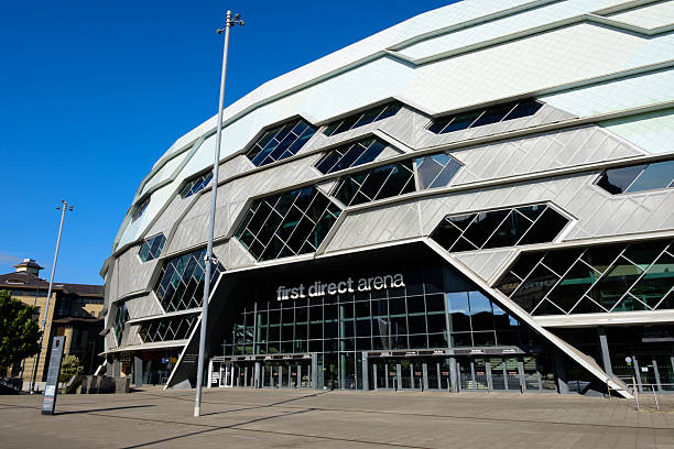 An exterior view of the Leeds First Direct Arena on a sunny day, showing the entrance with its distinctive geometric facade and large glass doors.