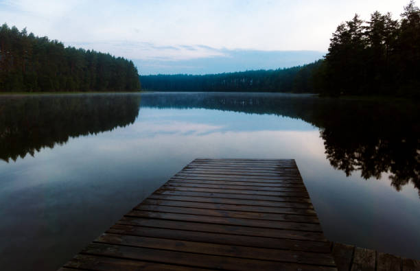 A wooden dock sits on the edge of a lake, with the water reflecting the sky above. The scene is peaceful and serene, with the calm water and the trees in the background creating a sense of tranquility