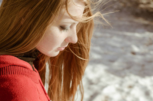 Young adult woman with long red hair gazes down, wearing red sweater. Sunlight highlights her features against blurred snowy background, creating serene winter atmosphere