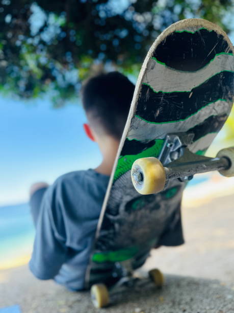 From a back-facing perspective, a teenage skateboarder sits near a sandy shoreline, the vibrant skateboard deck and wheels prominently in the foreground. The ocean glimmers in the background, capturing a carefree summer moment. Crisp lines of the board suggest active youth culture, while the relaxed posture conveys a brief pause to soak in the coastal scenery.