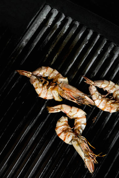 Three large prawns are being grilled on a black cast iron grill, displaying beautiful char marks from the heat. The seafood is cooked to perfection, showcasing its natural texture and caramelized surface. The dark background enhances the contrast, making the prawns the focal point of the composition. This image is perfect for seafood lovers, gourmet cooking enthusiasts, or restaurant menu designs featuring grilled seafood dishes.
