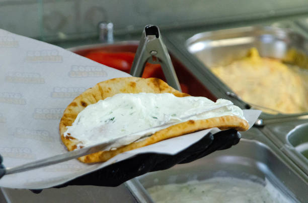 A man working at a food stand is spreading a creamy cucumber and sour cream sauce on a pita, preparing it for the other fresh ingredients. He carefully arranges the base, getting ready to add the perfectly grilled gyro and toppings.