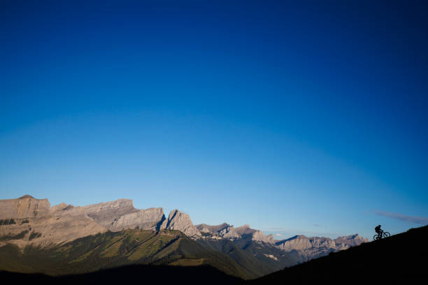 A man rides his e-bike mountain bike, or electric mountain bicycle, in the Rocky Mountains of Alberta, Canada, at sunrise. He rides a full-suspension, enduro-style mountain bike and wears a cycling helmet.