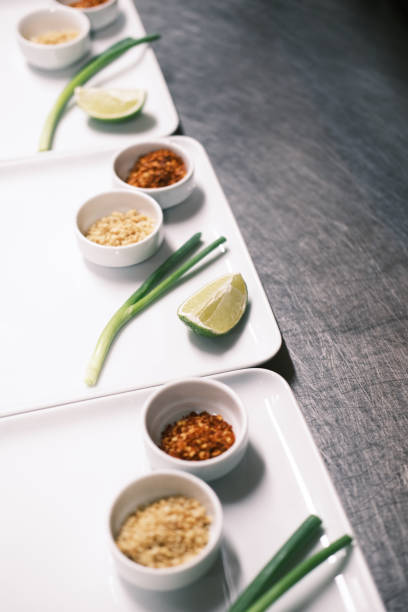 A visually appealing arrangement of essential Thai cooking ingredients on white plates, including crushed peanuts, chili flakes, lime wedges, and green onions. The minimalist presentation on a stainless steel countertop emphasizes the precision and artistry behind Thai cuisine. The clean and structured composition highlights the balance of flavors—spicy, sour, and nutty—integral to authentic Thai dishes. This image is perfect for food photography, restaurant branding, and content showcasing traditional Asian food preparation.