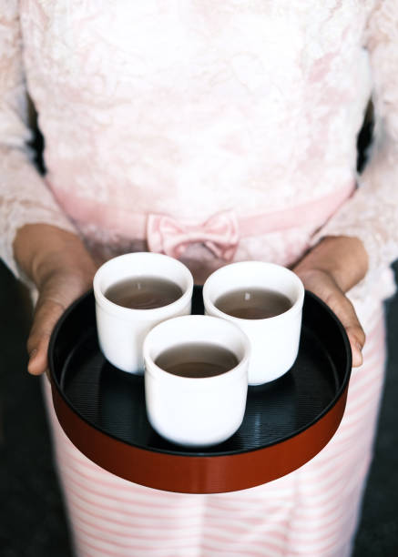 A close-up of a traditional tea-serving ceremony featuring three white cups filled with tea on a black lacquer tray. A woman dressed in elegant pink attire with delicate lace details and a bow holds the tray, symbolizing hospitality, mindfulness, and cultural traditions. The image reflects Asian tea culture, warmth, and the art of tea presentation.