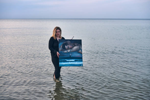 Young woman stands in calm ocean waters, holding surreal painting. Scene evokes tranquility and creativity, juxtaposing art with nature under softly lit sky