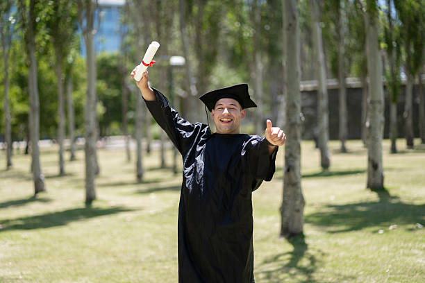 Cheerful young male wearing a red cape and graduation cap, holding a trophy while expressing excitement against a bright teal background.