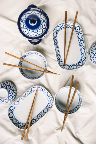 A beautifully arranged set of traditional Thai ceramic tableware featuring intricate blue and white patterns, accompanied by wooden chopsticks on a soft white fabric background. The elegant design and cultural significance of the dish set reflect Thai dining traditions, making this image perfect for use in restaurant branding, culinary photography, and cultural appreciation projects.
