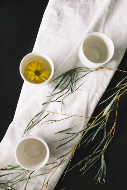 A serene tea composition featuring three white ceramic tea cups, one filled with herbal tea infused with a delicate yellow flower, placed on a soft linen fabric with dried leaves scattered around. This minimalist arrangement evokes a sense of tranquility and mindfulness, making it ideal for wellness concepts, tea culture, and natural lifestyle imagery.