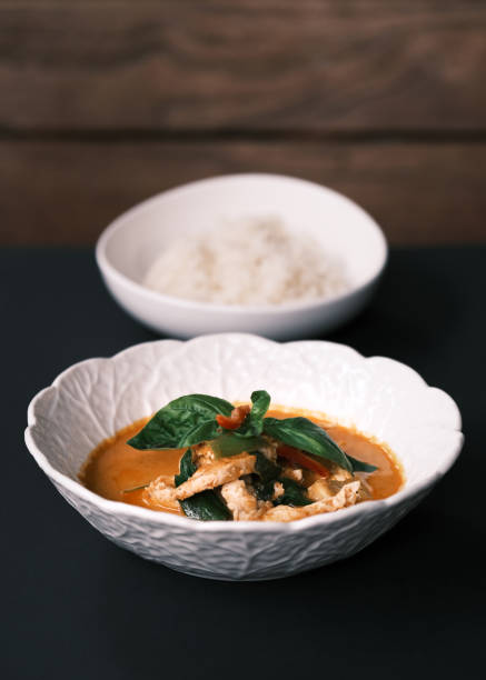 Close-up of Thai red curry with chicken and basil served in a white bowl, Traditional coconut milk-based curry with aromatic spices, Steamed rice in the background, Authentic Thai cuisine for restaurant menus and food photography