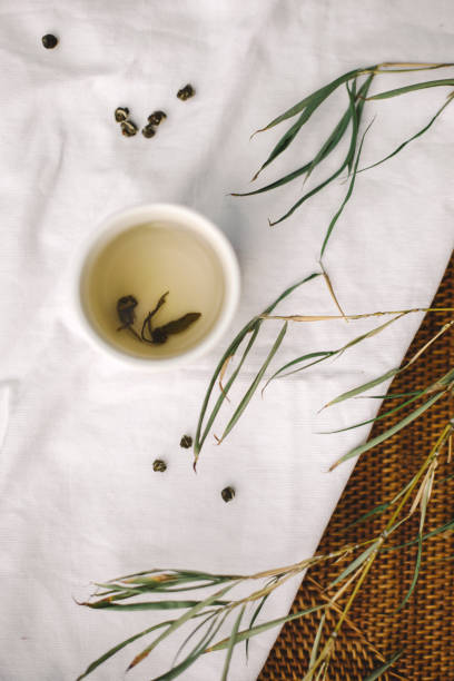 This image presents a delicate and artistic tea scene, featuring a white ceramic cup filled with freshly brewed green tea. Floating tea leaves and scattered loose tea pearls enhance the organic and natural aesthetic. The soft linen background, combined with delicate plant elements and woven textures, creates a peaceful and minimalist tea ceremony ambiance. Ideal for tea branding, wellness concepts, and mindful living photography, evoking a sense of tranquility and relaxation.