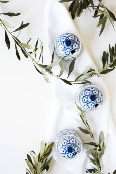 A beautifully styled composition featuring three blue and white ceramic bowls placed on soft white fabric, accompanied by fresh olive branches. The arrangement highlights traditional Asian tableware with a touch of Mediterranean influence, creating a blend of cultural aesthetics. The minimalist and elegant presentation is ideal for culinary themes, restaurant branding, food photography, and interior decor concepts.