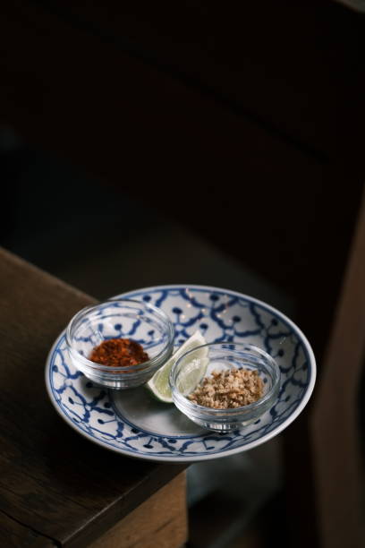 A beautifully arranged set of traditional Thai condiments featuring chili flakes, crushed peanuts, and a lime wedge, elegantly placed in small glass bowls on a classic blue and white ceramic plate. The dark and moody background enhances the rustic authenticity of Thai cuisine, emphasizing the balance of flavors essential in Thai cooking. The intricate patterns of the plate add cultural depth, making this image perfect for food photography, restaurant branding, and content celebrating traditional Asian culinary elements.