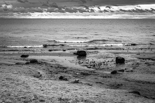 A bold and dramatic black and white seascape featuring ocean waves rolling onto a textured sandy shoreline with scattered rocks. The moody sky, filled with layered clouds, enhances the depth and contrast of this coastal landscape. This high-contrast monochrome image evokes a sense of solitude and tranquility, making it ideal for editorial use, travel photography, environmental themes, and minimalist fine art prints. Perfect for designers, publishers, and content creators seeking atmospheric ocean imagery