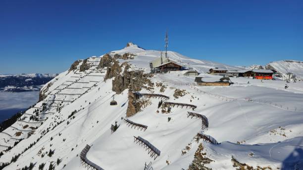 istock A scenic mountaintop resort sits atop a snow-covered peak, with a cable car ascending the rugged slopes. Wind barriers line the hillside, while a communications tower stands against the clear blue sky. 2201973693