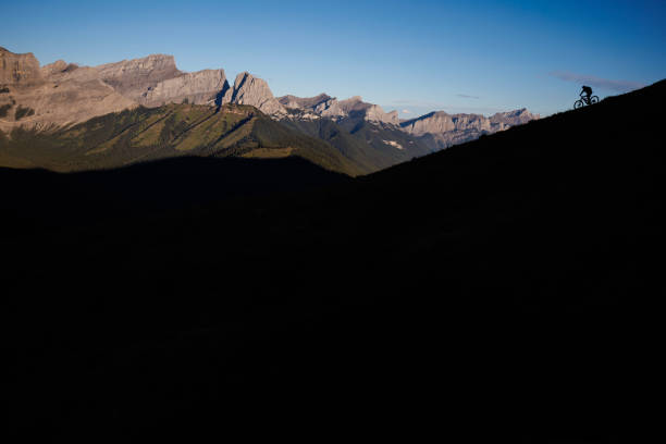 A man rides his electric mountain bike down a hill on a trail in the Rocky Mountains of Alberta, Canada, at sunrise. He rides a full-suspension, enduro-style e-mountain bike and wears a cycling helmet.