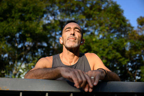 Mature hispanic athlete resting in a park after exercising, leaning against a fence while enjoying the tranquility of nature, wearing wireless earphones and soaking up the summer sun