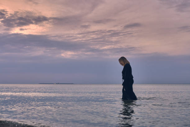 Young woman in black dress wades through calm ocean waters during tranquil sunset. Pink hues of sky evoke sense of solitude and peace, symbolizing introspection
