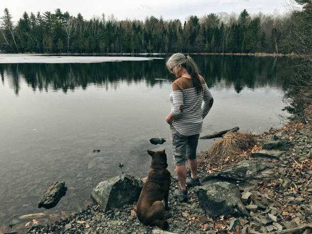 Fit senior woman, a behind view, on a hike with her dog, pausing by the lake and looking down at her dog, enjoying the water views and surrounding forest. Taken in Northern Wisconsin.