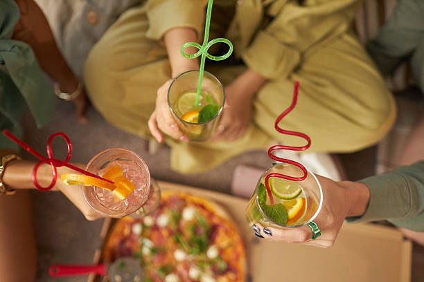 High angle view of unrecognizable three girls sitting at coffee table with pizza on it, holding glasses with soft drinks